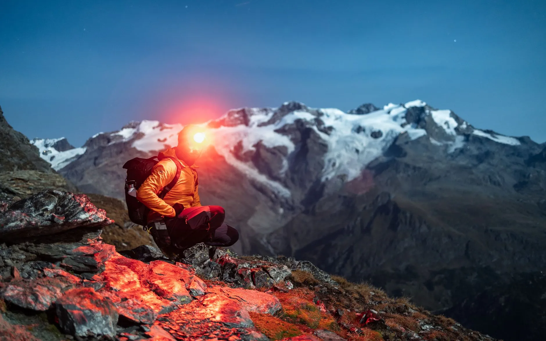 Person in oranger Jacke sitzt in der Daemmerung auf Fels, der Lichtkegel der Stirnlampe zeigt zum verschneiten Berg
