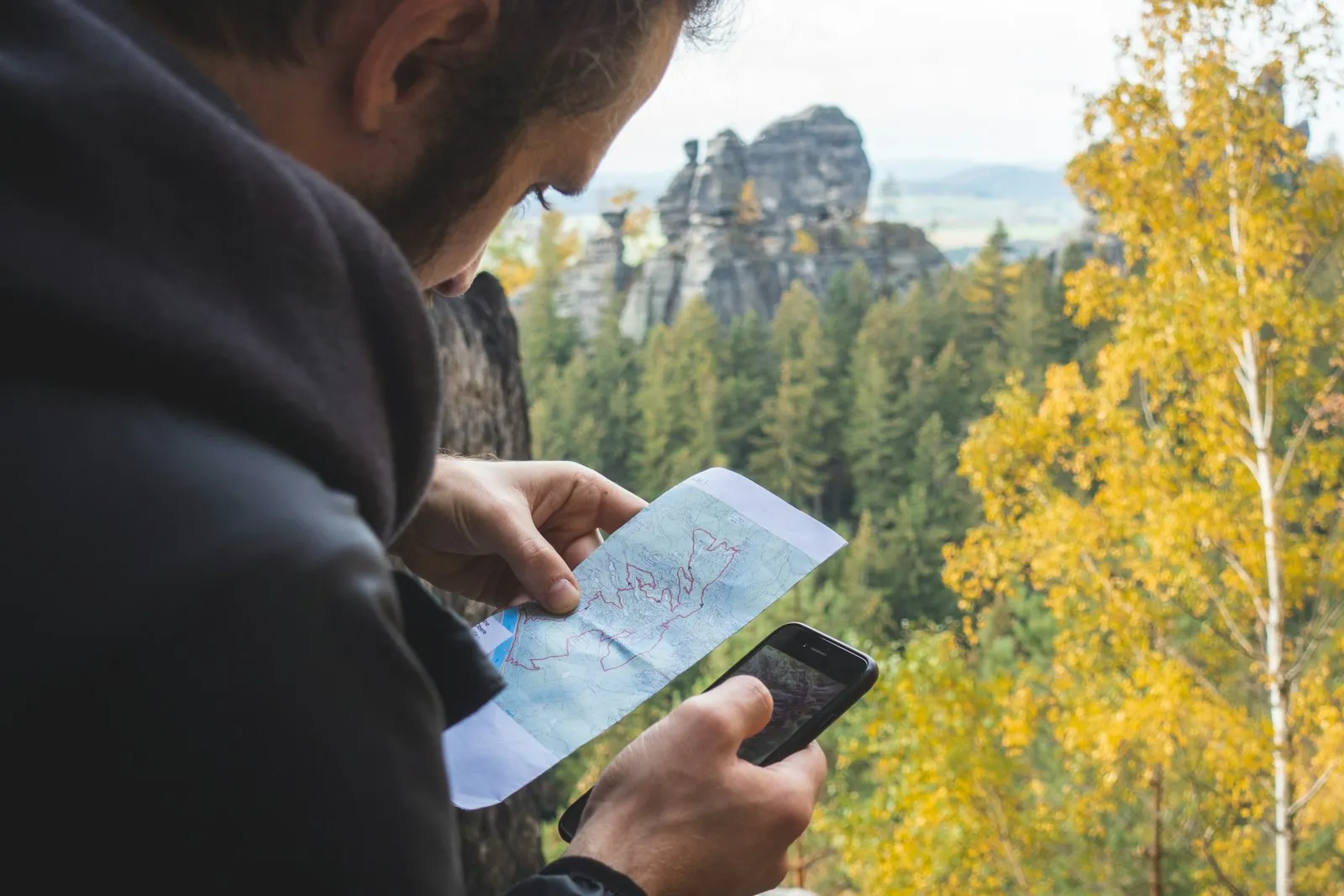 Person haelt Papierkarte und Smartphone nebeneinander, dahinter Sandsteinfelsen und Herbstwald