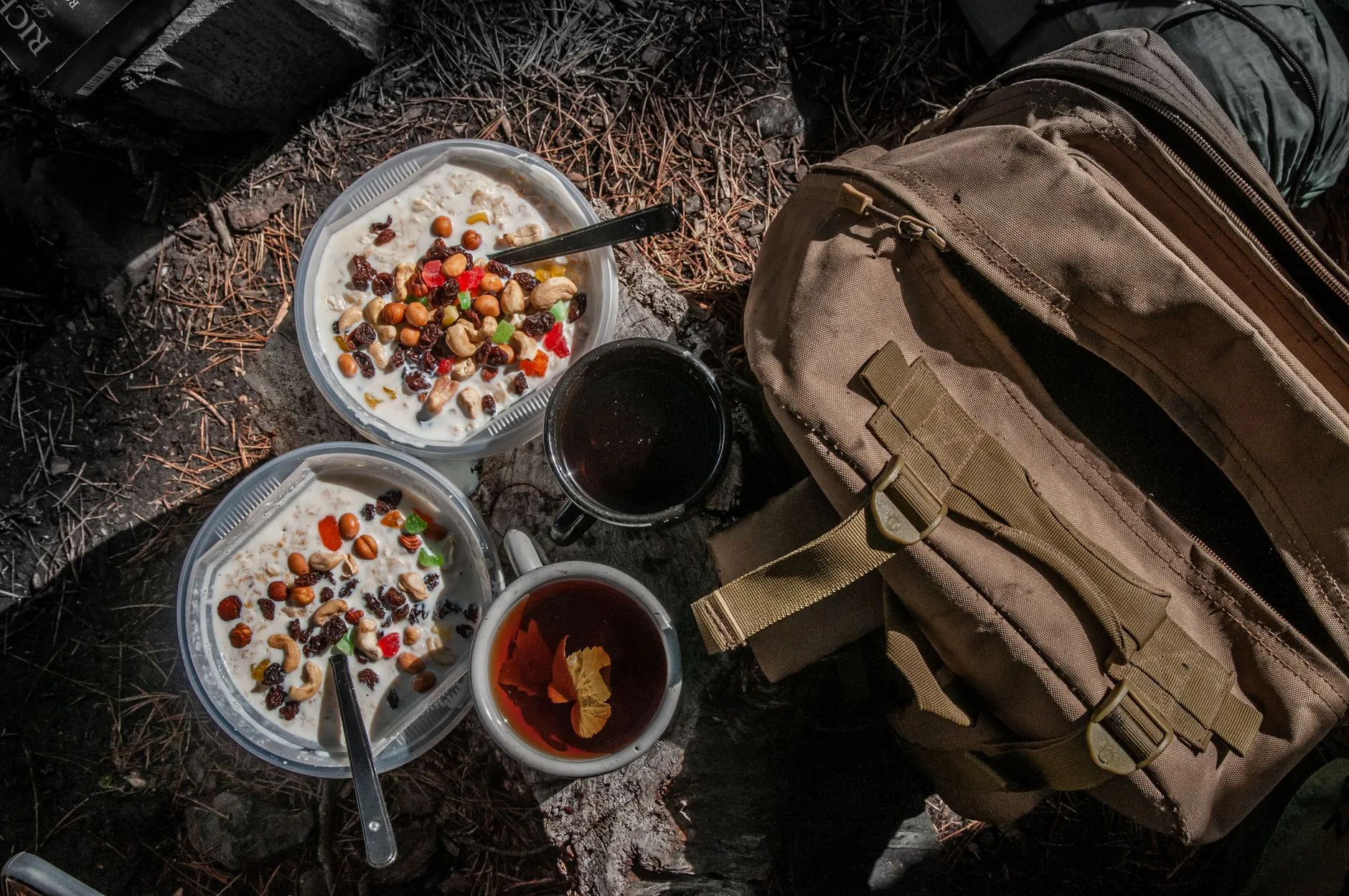 Zwei Schalen Müsli mit Nüssen und Beeren, daneben zwei Becher Tee und ein Rucksack auf Waldboden