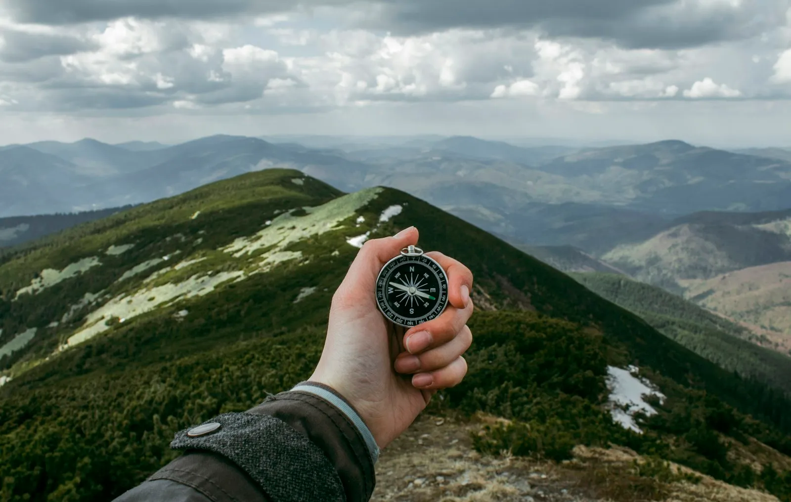 Ausgestreckte Hand haelt einen Kompass ueber einen Bergruecken, darunter Taeler und Wolken