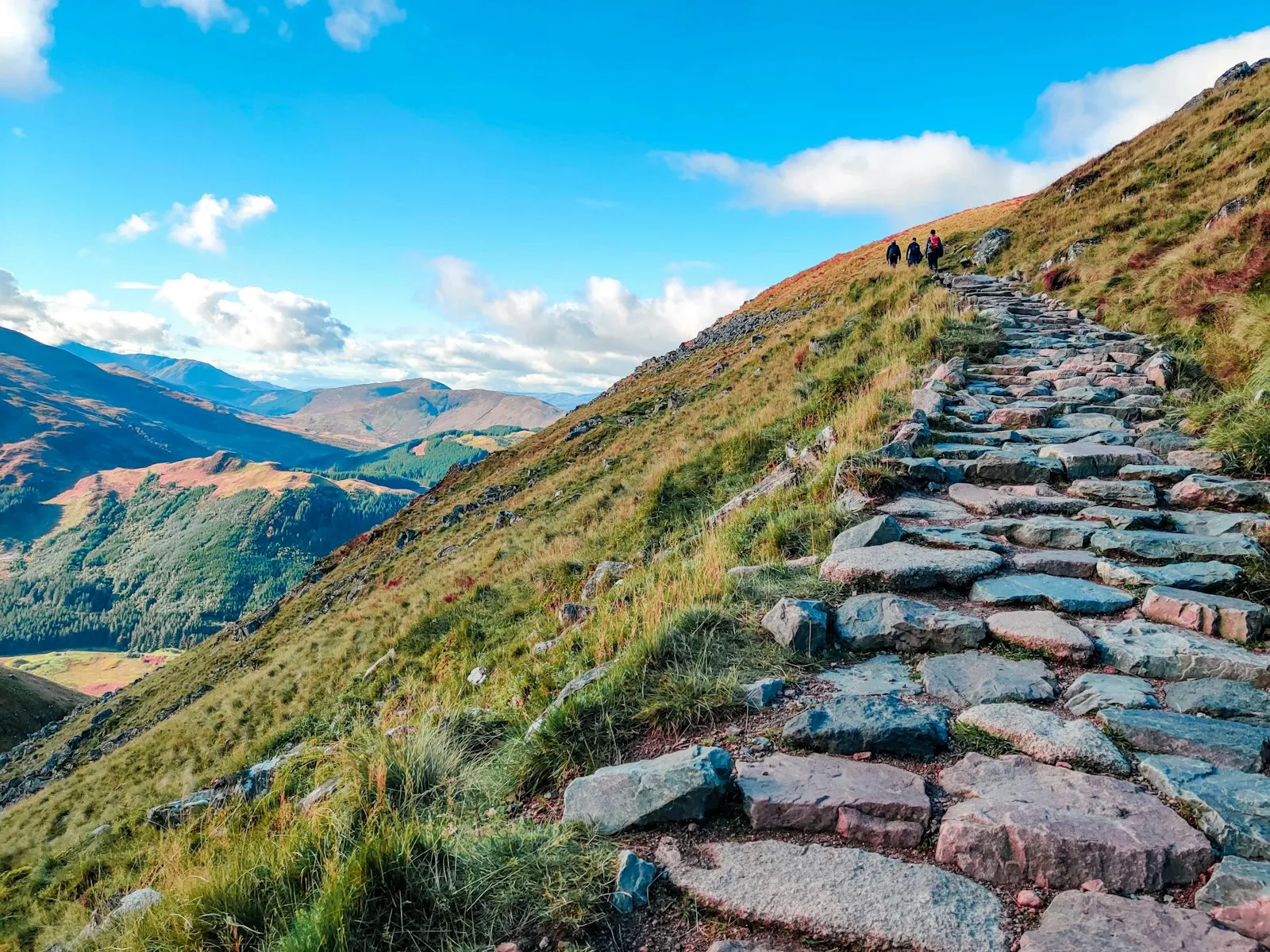 Gepflasterter Steig quert einen Grashang, in der Ferne zwei Wanderer und ein weites Tal