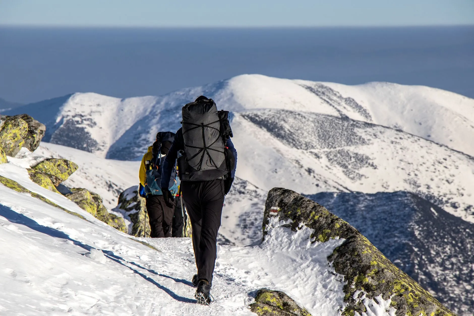 Zwei Personen mit grossen Rucksaecken gehen auf einem Pfad ueber einen schneebedeckten Bergruecken