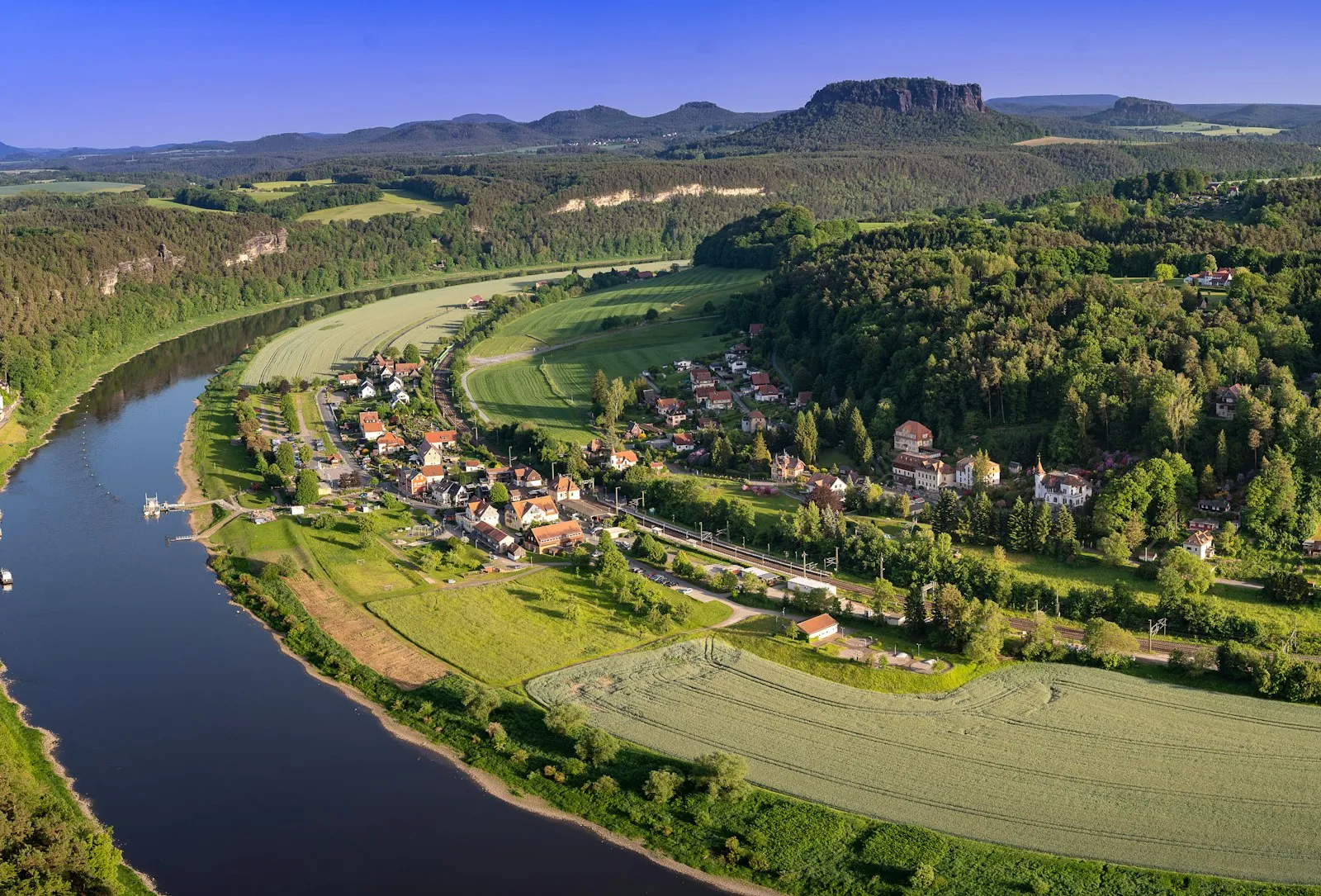 Luftbild eines Elbbogens mit Ortslage, Feldern und Wald, im Hintergrund ein Tafelberg