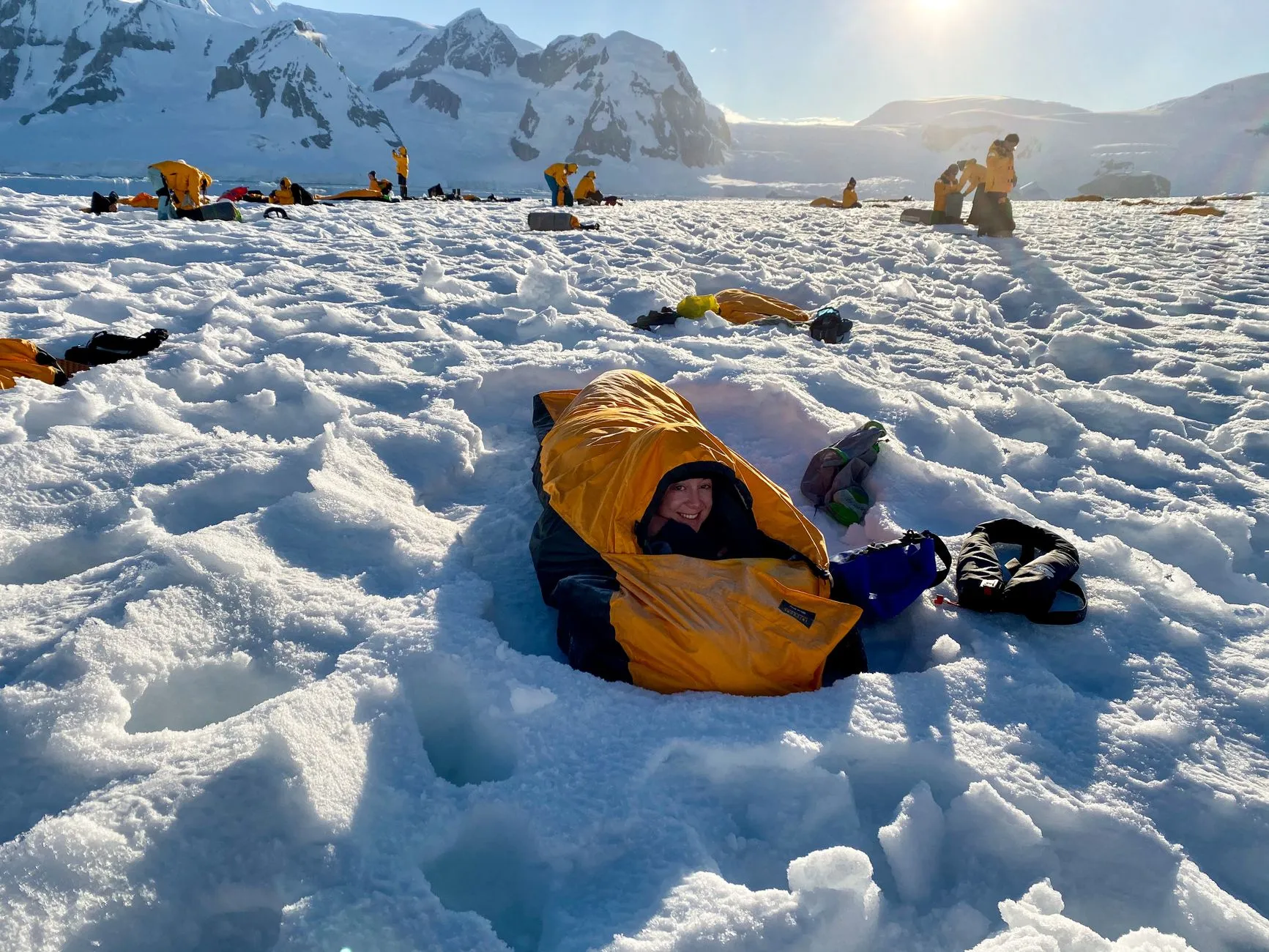 Person liegt in einem orangefarbenen Schlafsack direkt im Schnee, dahinter ein Lager vor verschneiten Bergen