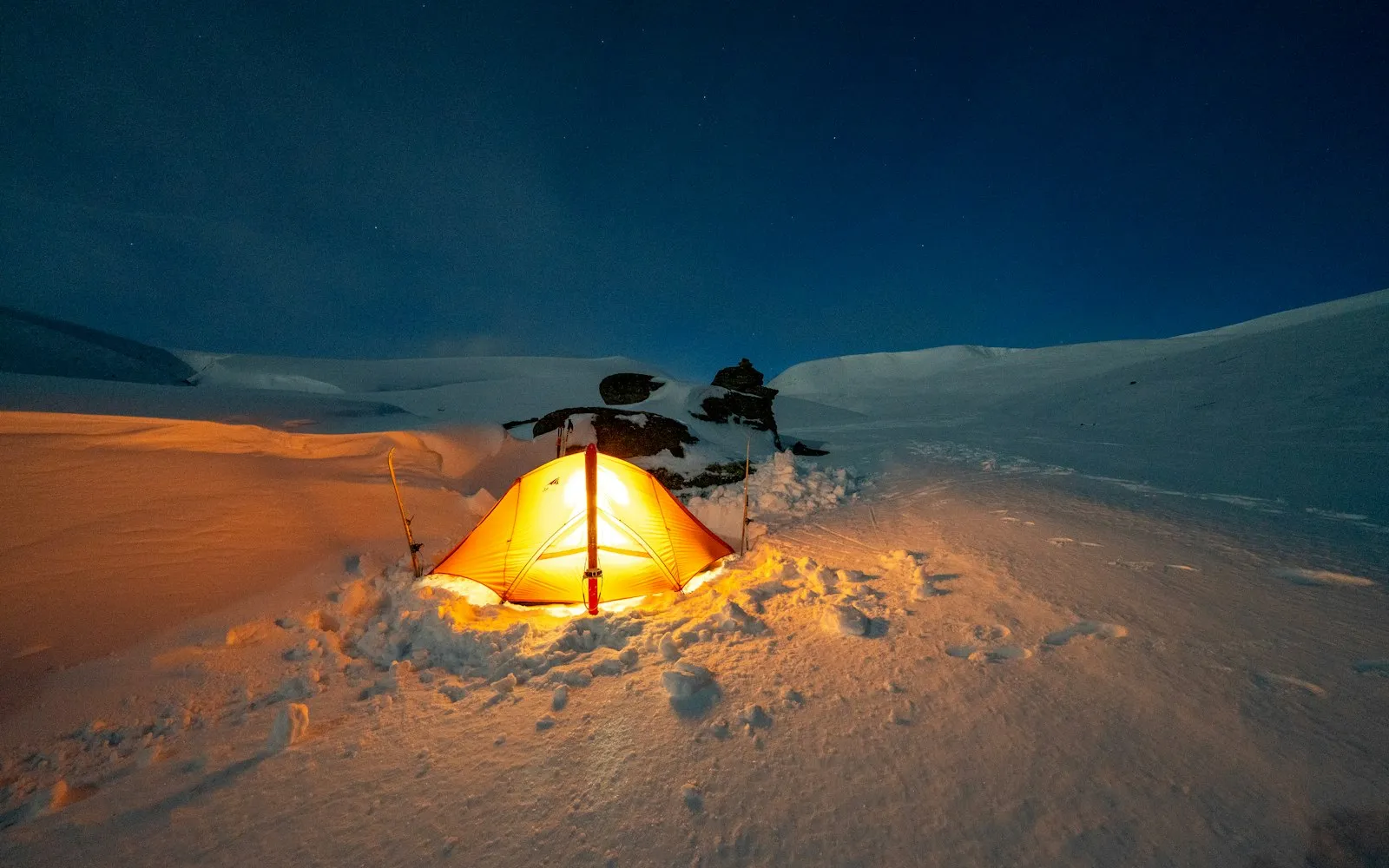Von innen beleuchtetes Zelt nachts in einer verschneiten Landschaft unter blauem Himmel