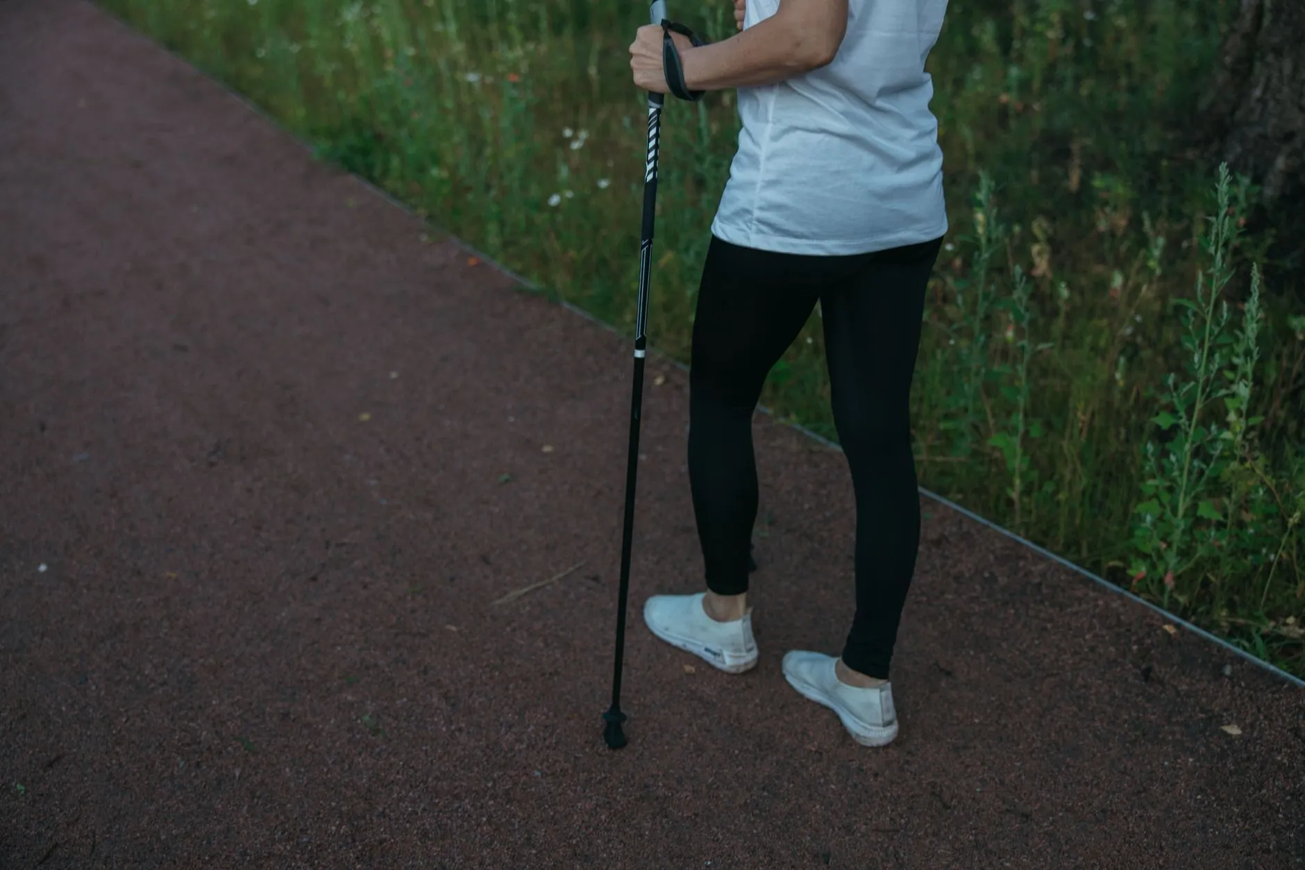 Person steht auf einem Weg und haelt einen Trekkingstock senkrecht neben sich, Blick auf Hand und Beine