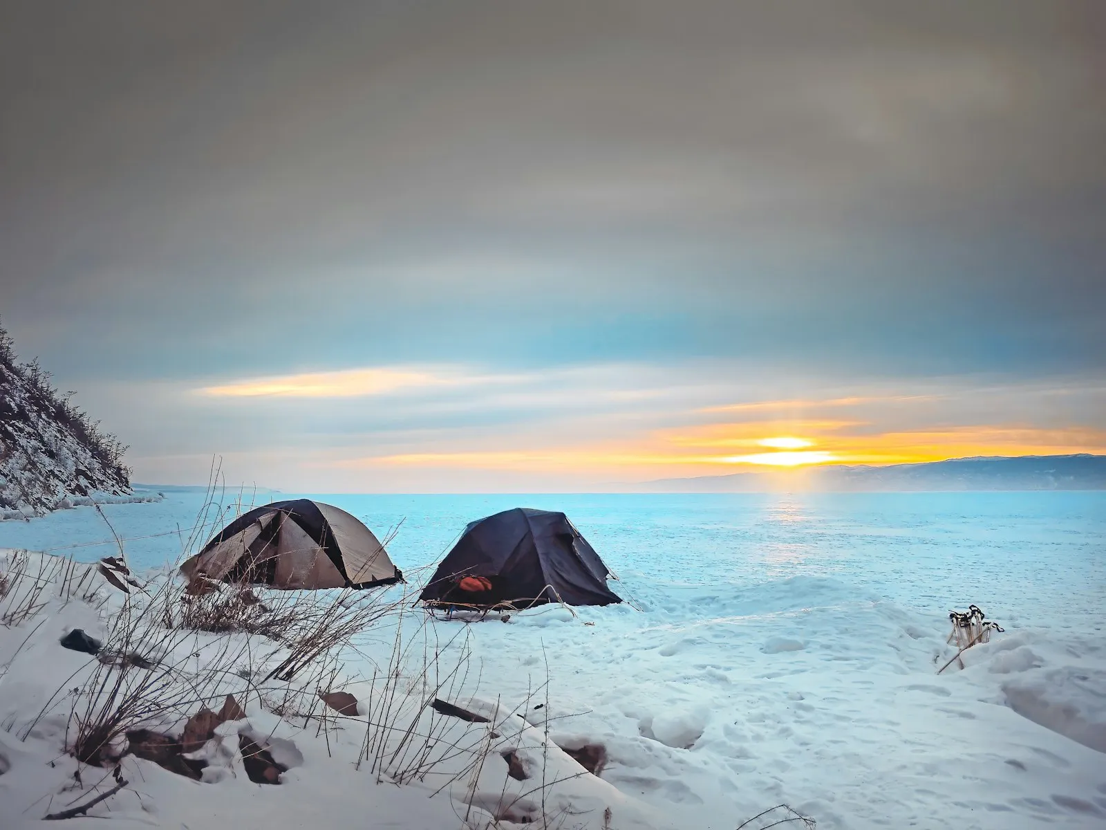 Zwei kleine Zelte stehen auf einer verschneiten Flaeche vor einem Gewaesser bei Sonnenuntergang