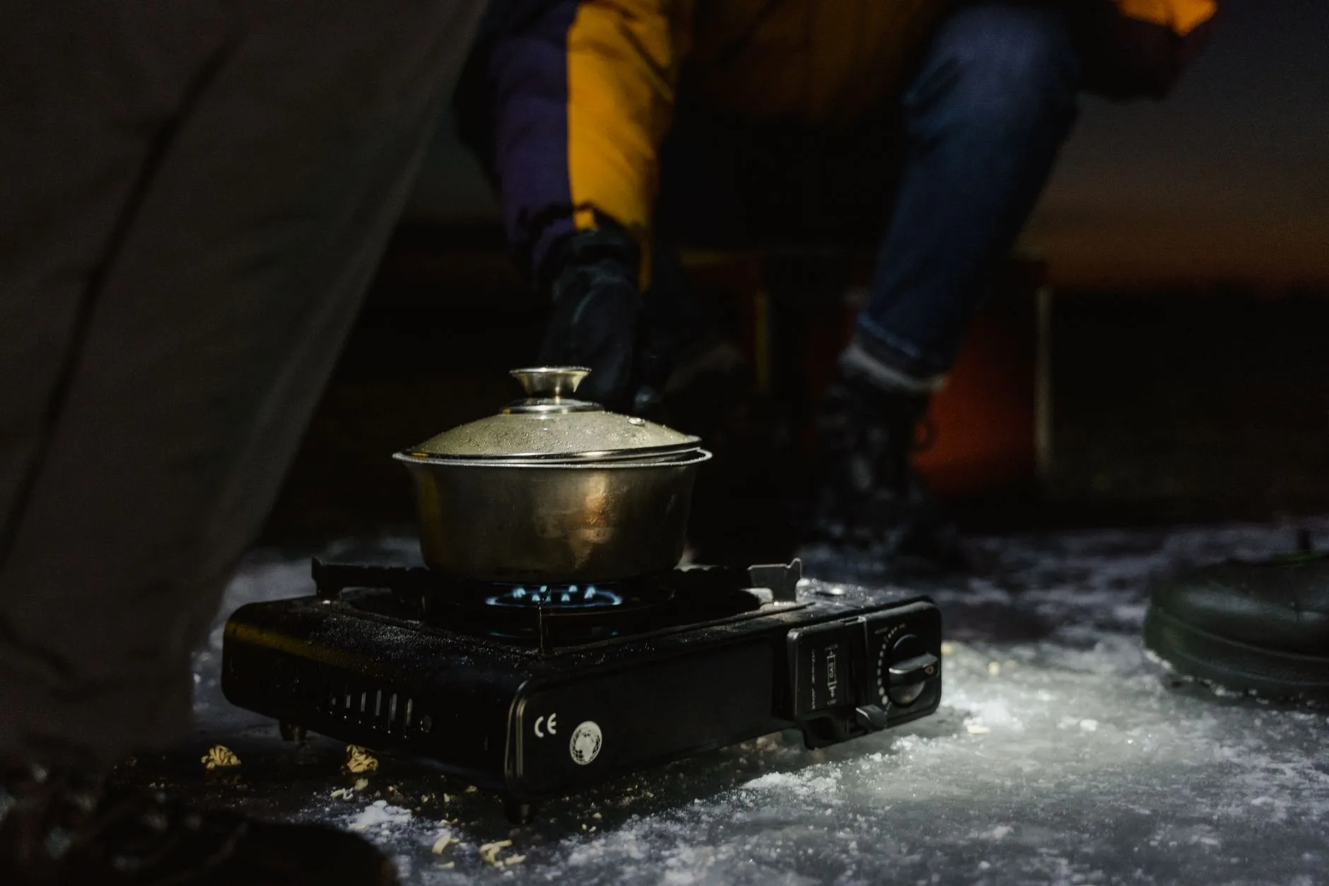 Topf mit Deckel auf einem Gaskocher, der nachts unmittelbar auf einer Schneeflaeche steht, Flamme sichtbar